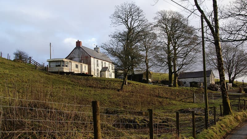 White-painted hill farmhouse and outbuildings on a sloping field with bare trees and a wire fence in rural Wales